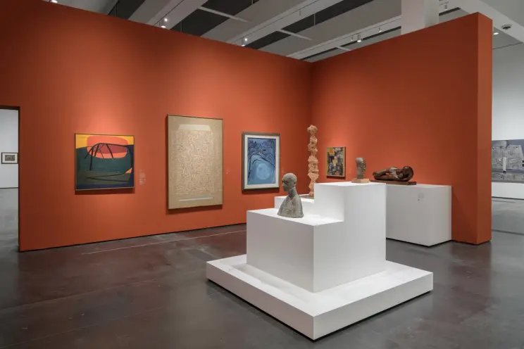 An art gallery room with various art pieces displayed against an orange exhibition wall. Additional pieces are presented o...