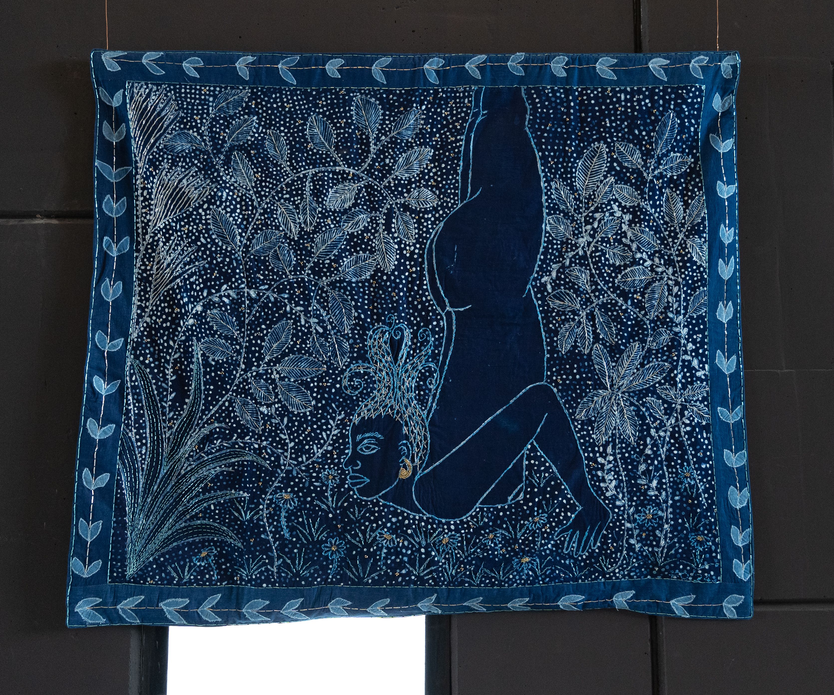A vibrant indigo textile depicting a human figure amid detailed botanical patterns.