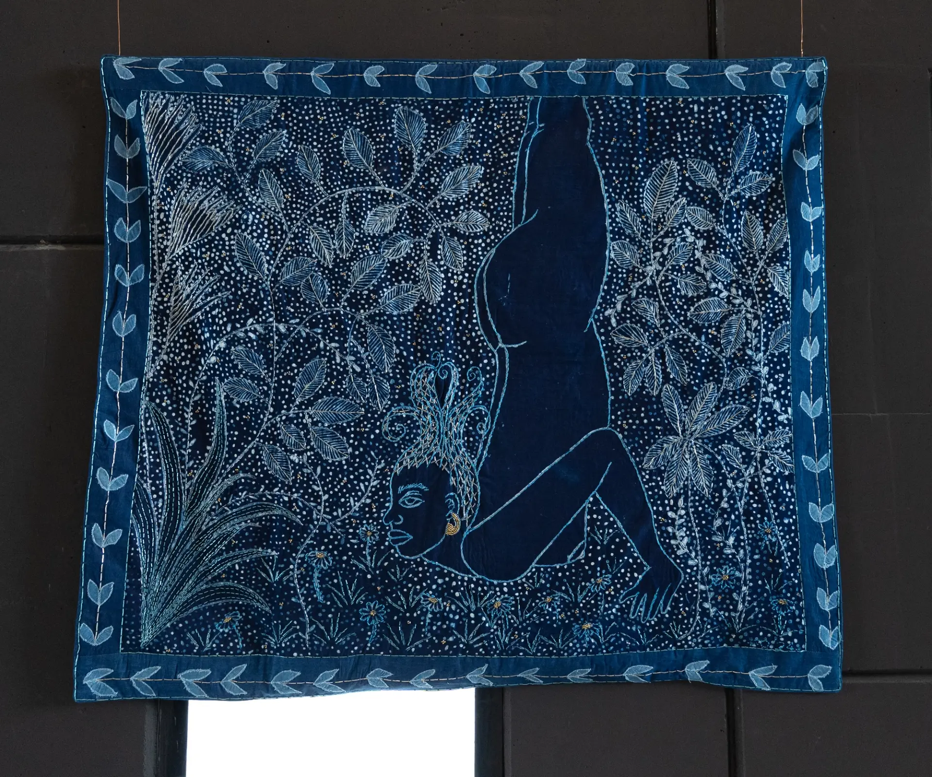 A vibrant indigo textile depicting a human figure amid detailed botanical patterns.
