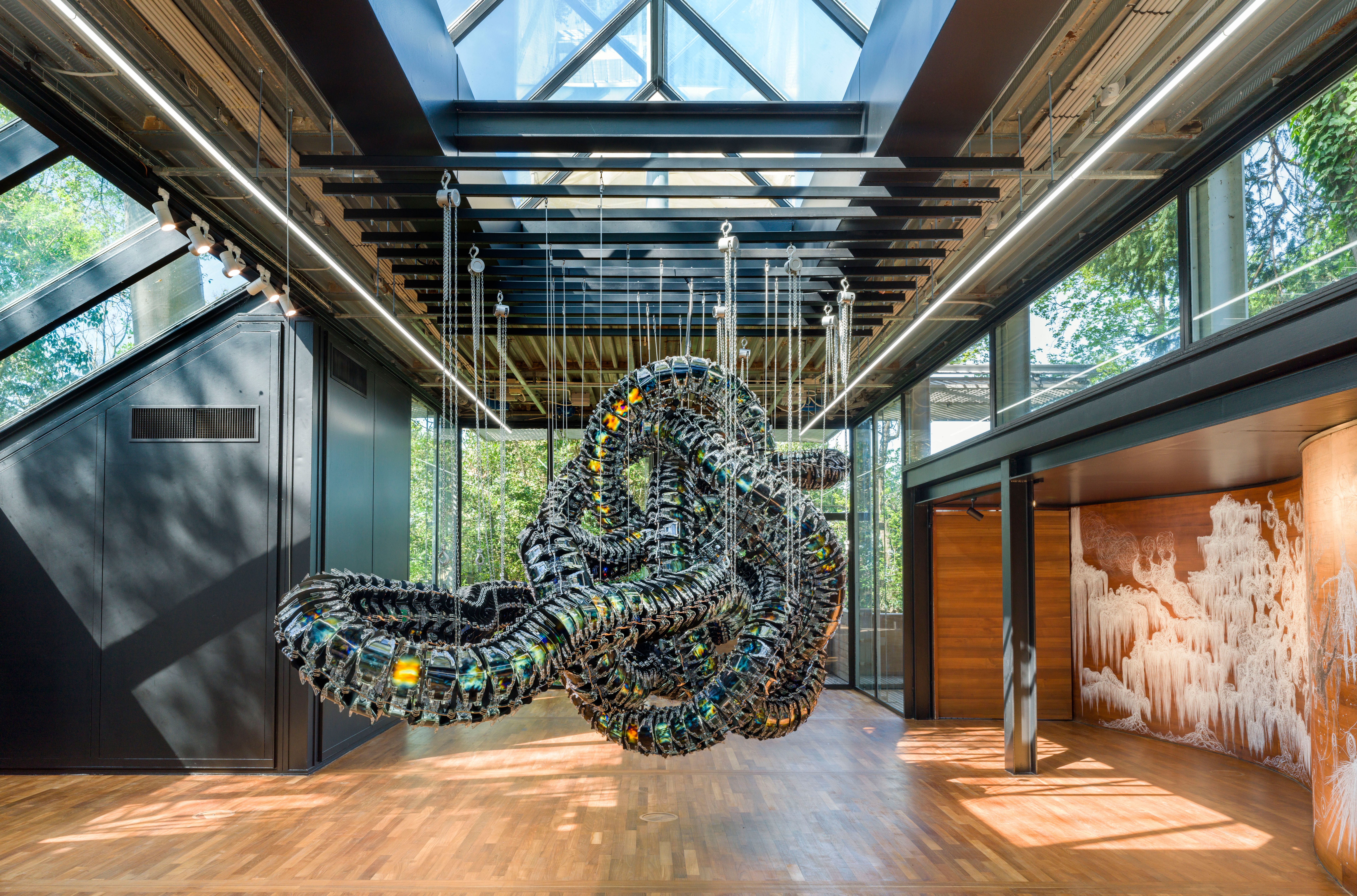 A metallic art installation reminiscent of a snake node is suspended from the ceiling in a gallery hall, surrounded by oth...