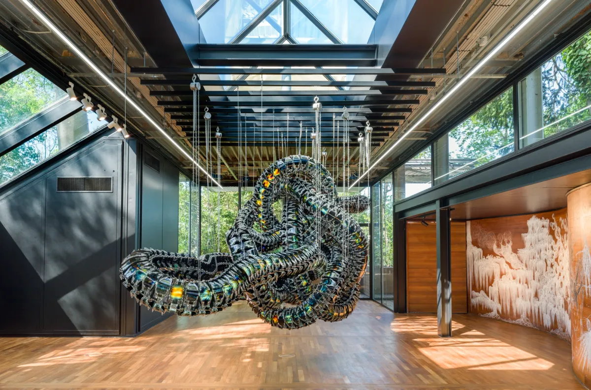 A metallic art installation reminiscent of a snake node is suspended from the ceiling in a gallery hall, surrounded by other diverse art pieces.
