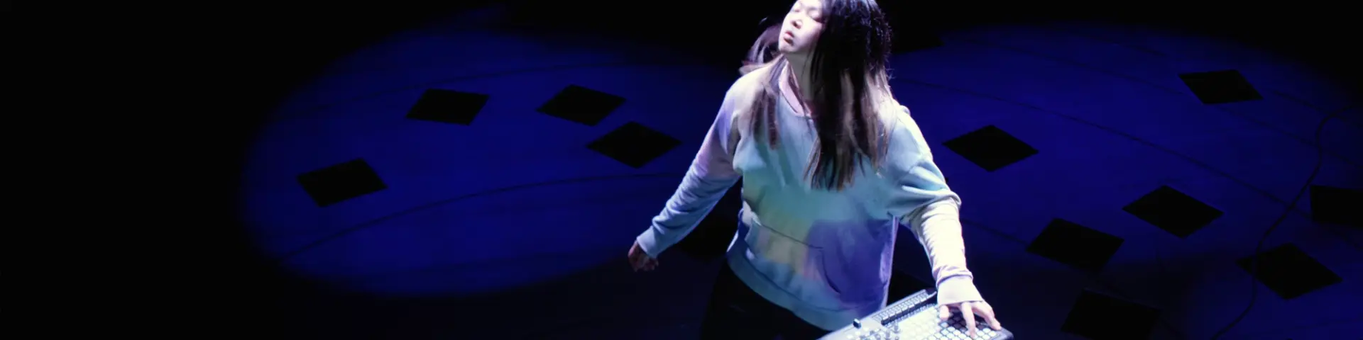 A human figure stands on a stage, engaging in a performance using a midi controller for an audiovisual installation, under...