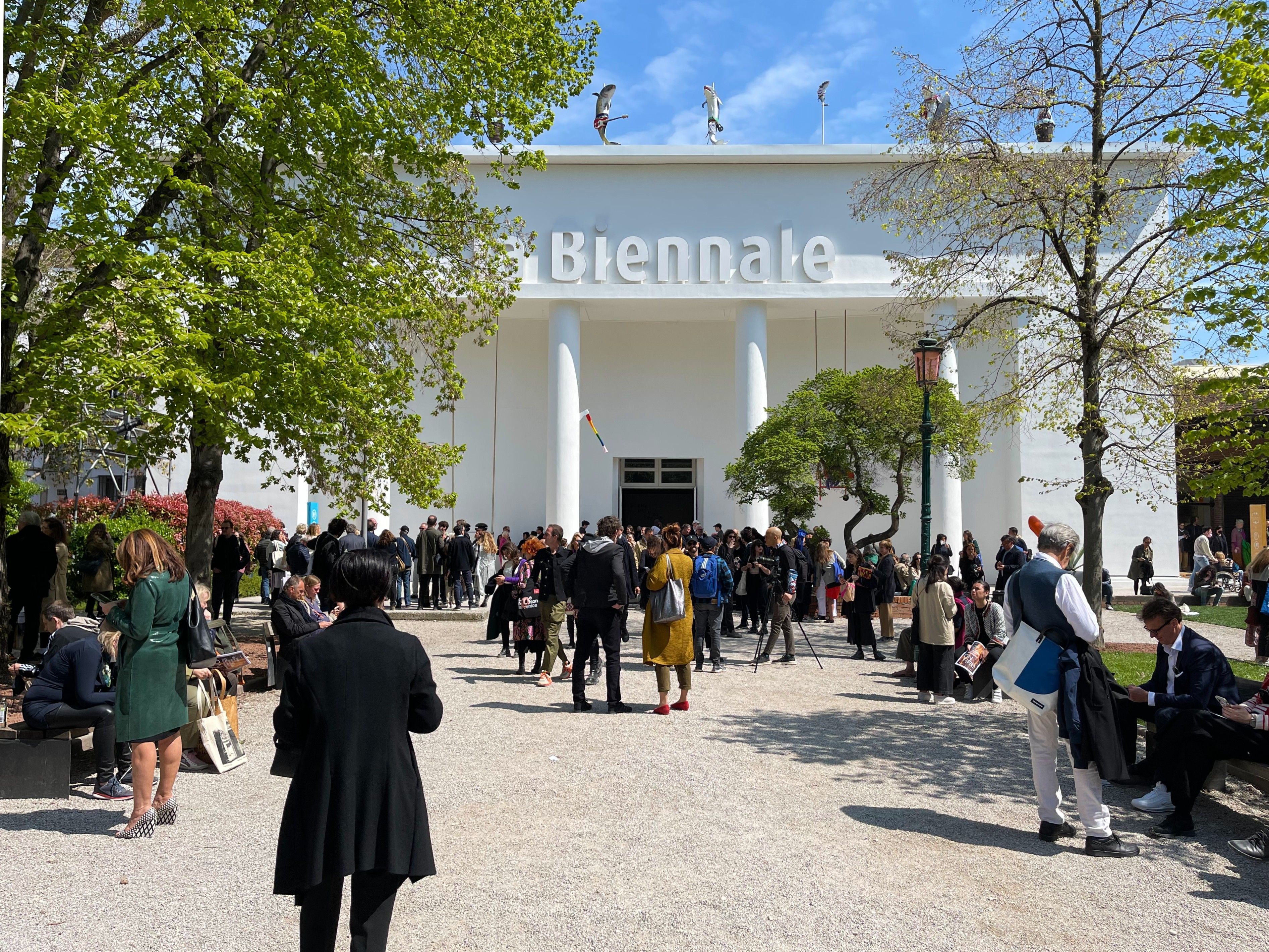 People gathered outside a white building under a clear sky surrounded by trees.