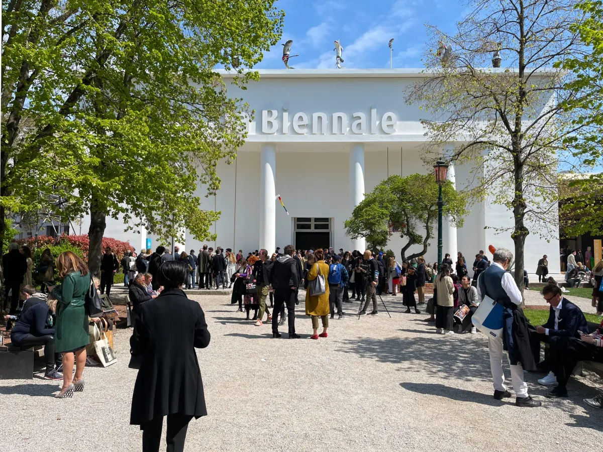 People gathered outside a white building under a clear sky surrounded by trees.