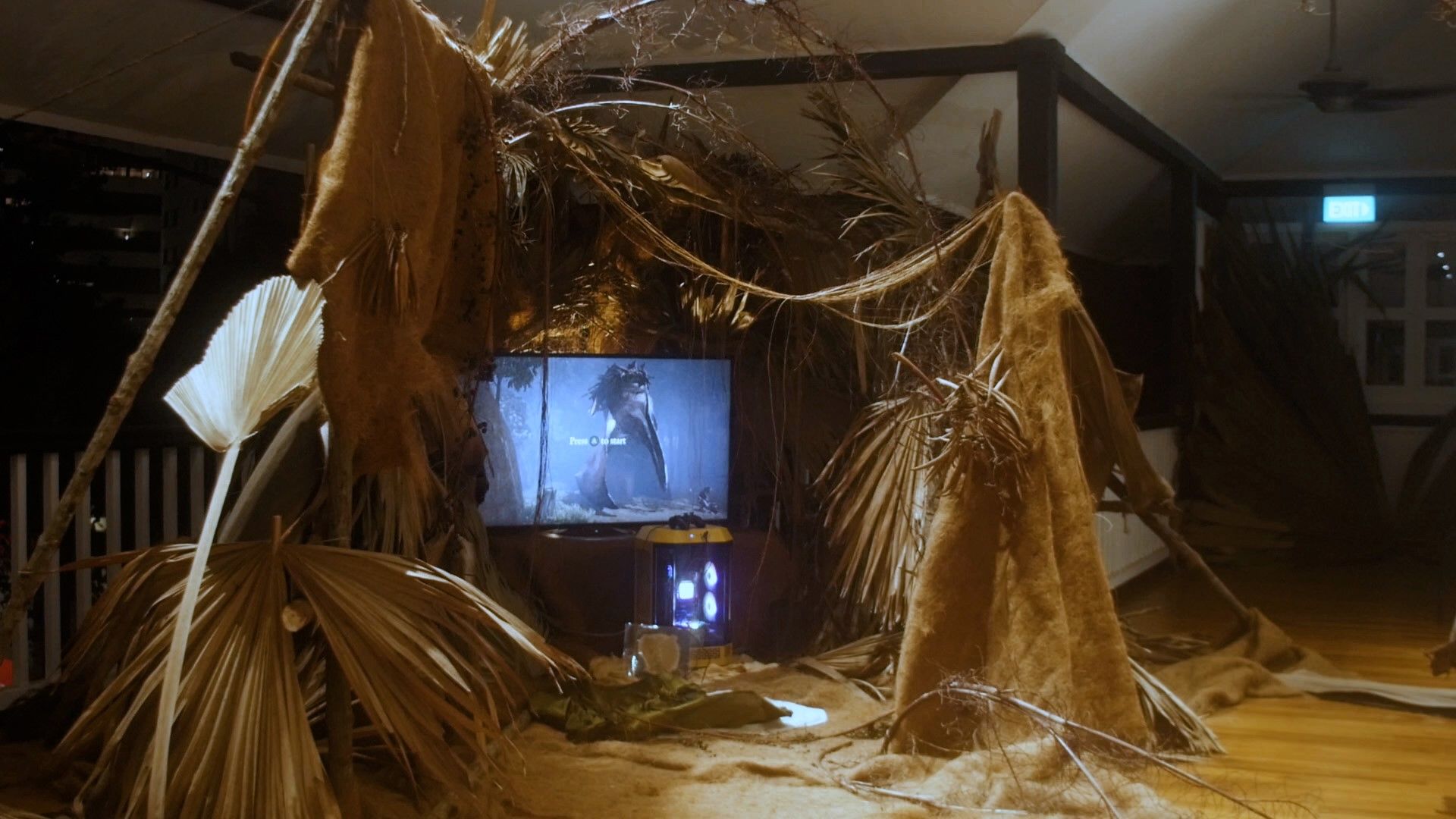 An installation featuring natural materials surrounding a gaming setup displaying a video game screen.