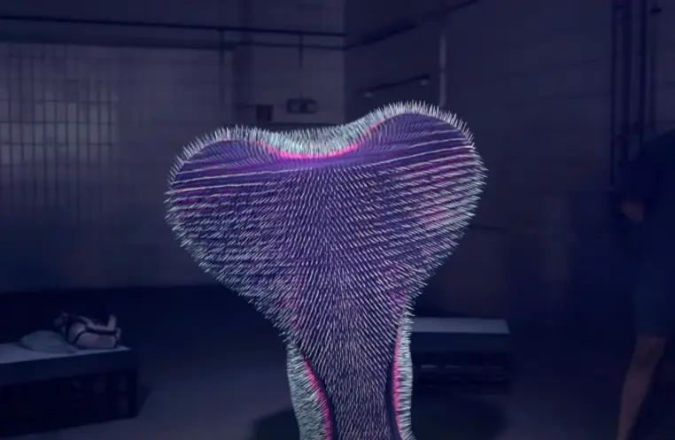 A spiky, heart-like form illuminated with purple and magenta light sits in the center of a dimly lit room. The room has wh...