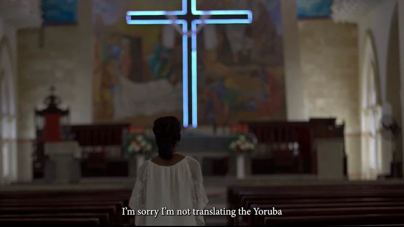 A woman stands alone in a church, her back to the camera, in front of a glowing blue cross.