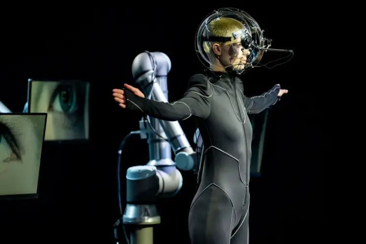 A person in a futuristic suit and helmet stands with robotic arms in the background, presenting an innovative technology and fashion integration.