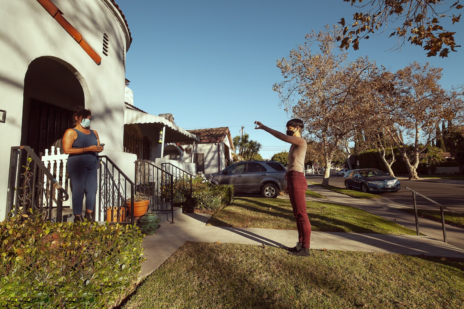 An individual in a mask stands near an autumnal building, showcasing an object in their hand to another person standing on...