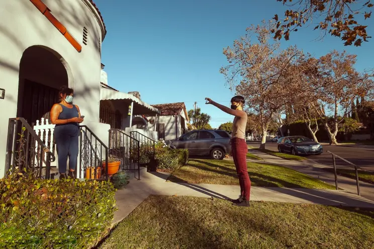 An individual in a mask stands near an autumnal building, showcasing an object in their hand to another person standing on...