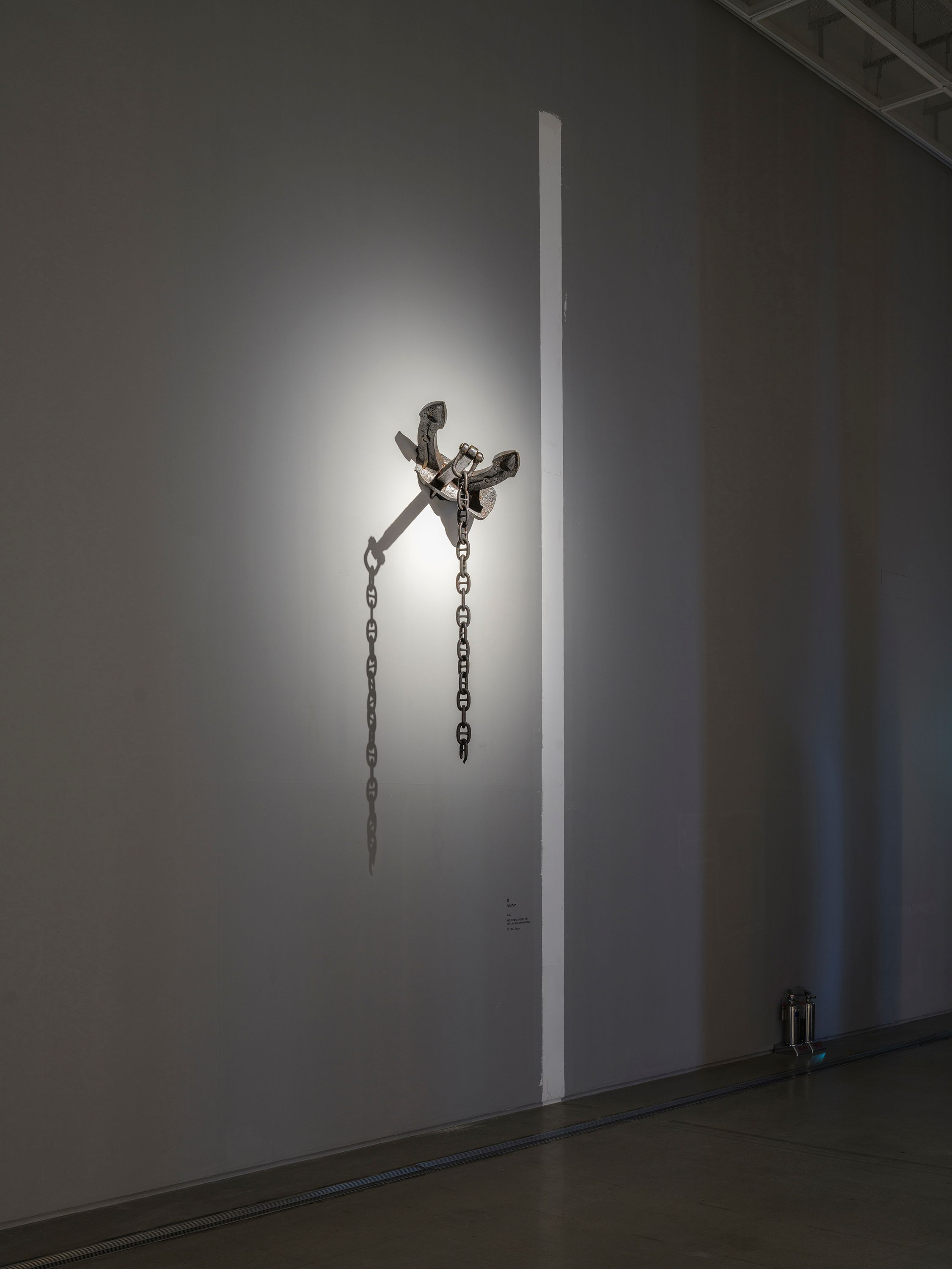 An anchored wall under a spotlight, with a broken chain hanging from the anchor, viewed in a dimmed environment.