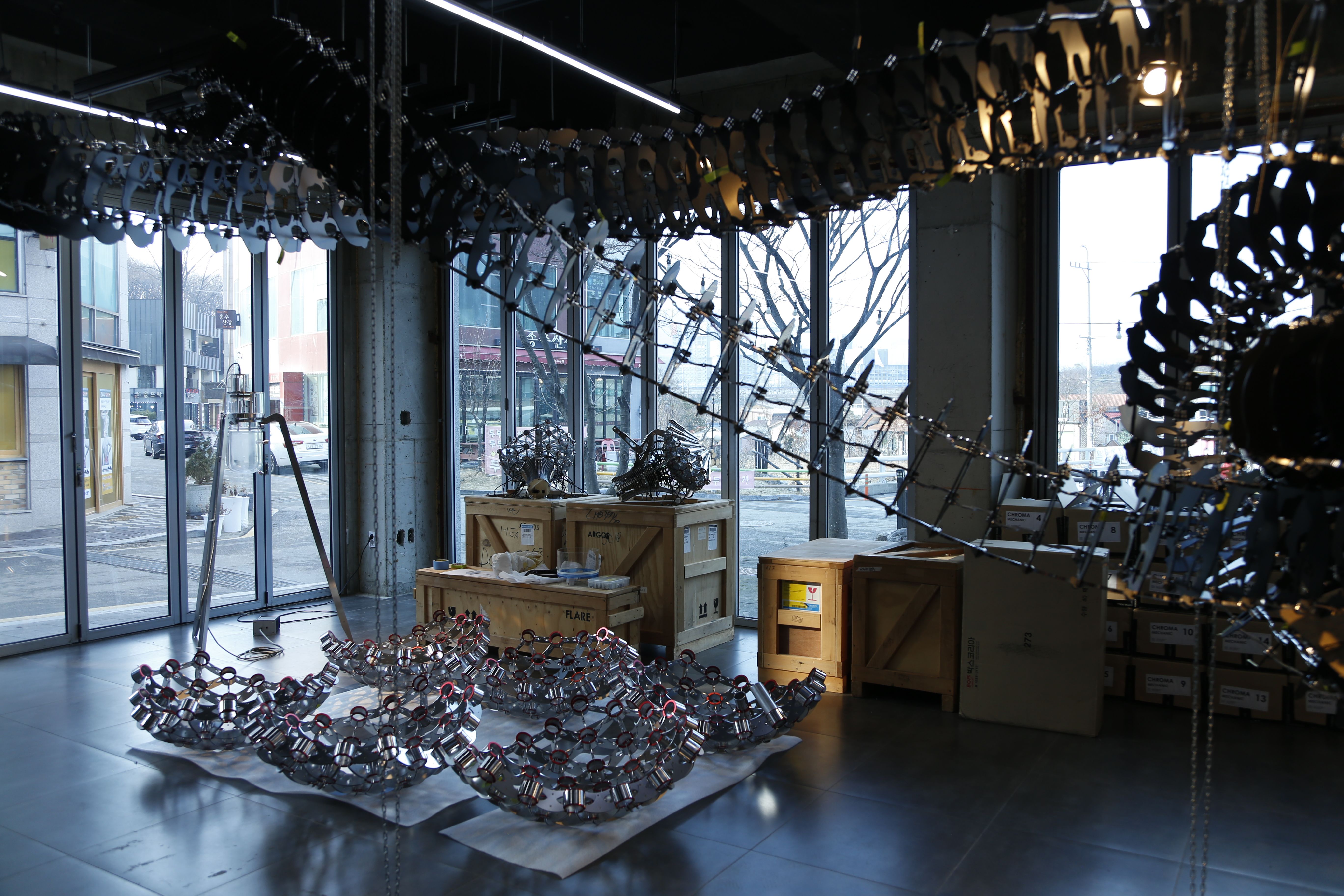 A room with windows houses several metallic pieces, awaiting assembly in a gallery space.