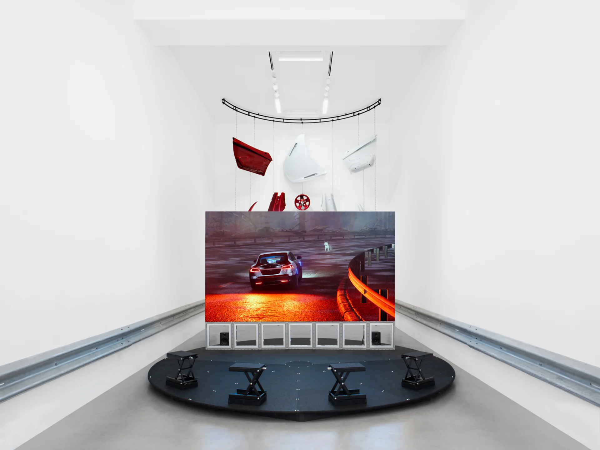 A minimalist white gallery featuring a video of a car on a dark, rainy road surrounded by suspended auto body parts.