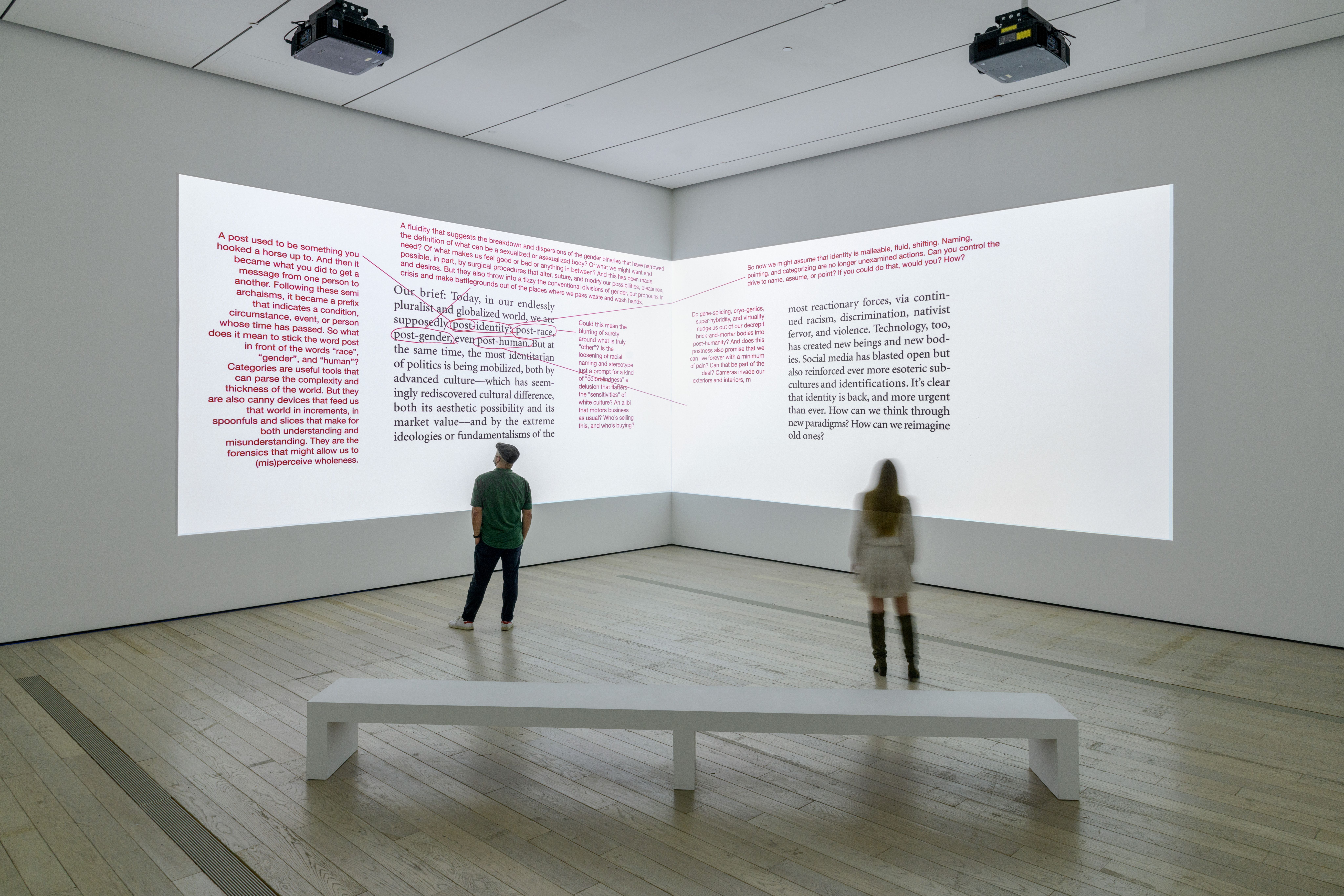 Two individuals stand in a gallery space, observing projected text media with red corrections on dual walls. A solitary wh...