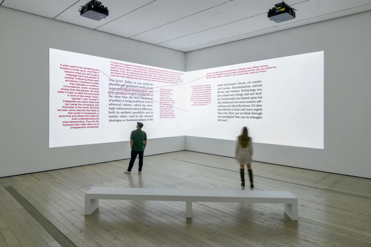 Two individuals stand in a gallery space, observing projected text media with red corrections on dual walls. A solitary white bench sits in the foreground.