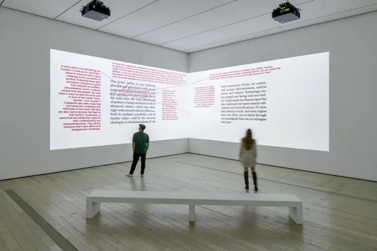 Two individuals stand in a gallery space, observing projected text media with red corrections on dual walls. A solitary wh...