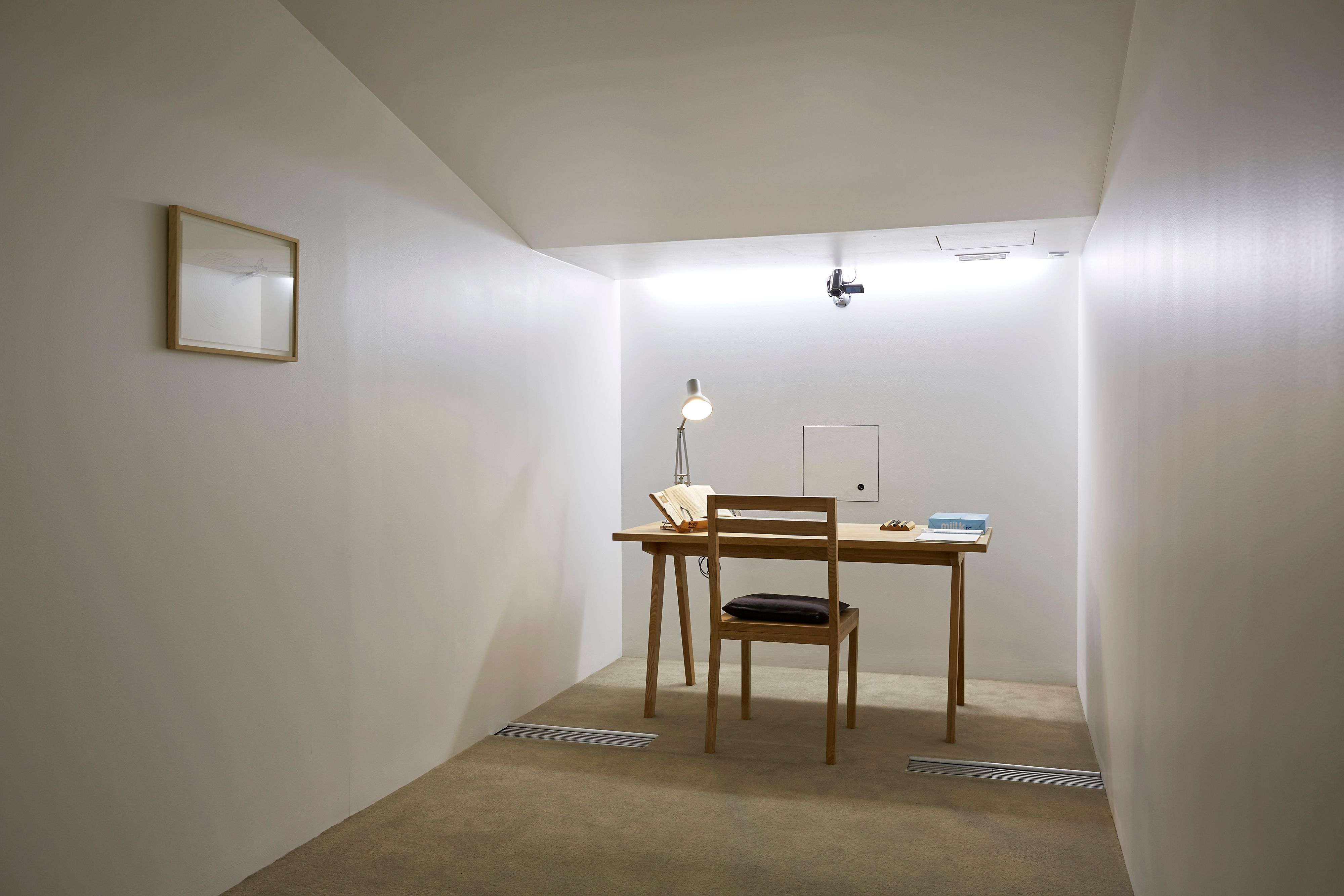 A narrow, white room featuring a wooden desk cluttered with books and a lamp, next to a wooden chair. An image frame hangs...