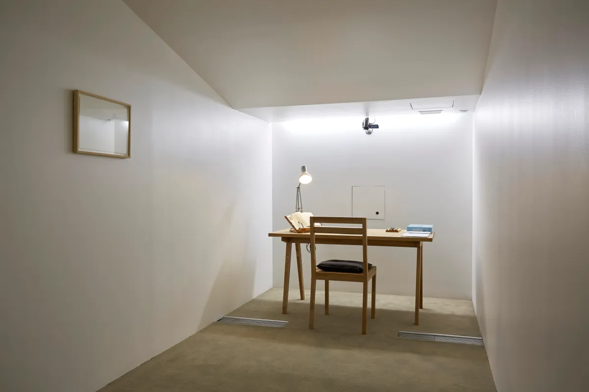 A narrow, white room featuring a wooden desk cluttered with books and a lamp, next to a wooden chair. An image frame hangs on the side wall.