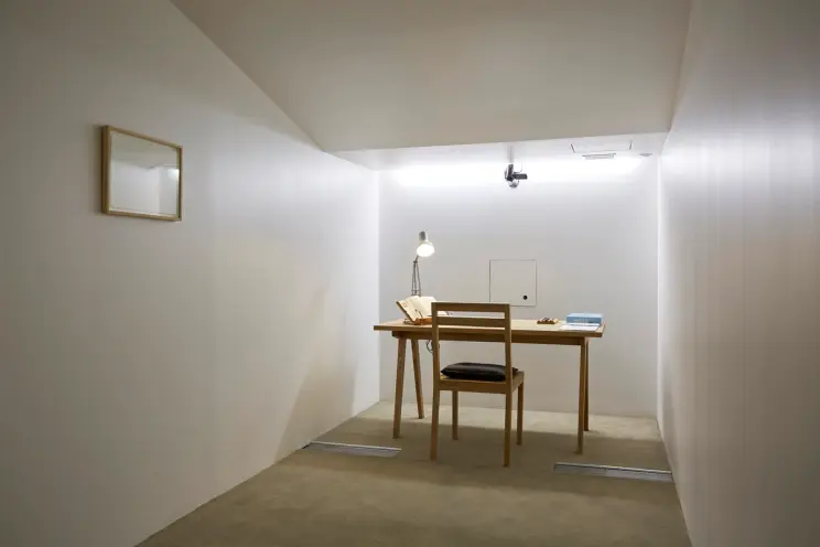 A narrow, white room featuring a wooden desk cluttered with books and a lamp, next to a wooden chair. An image frame hangs...