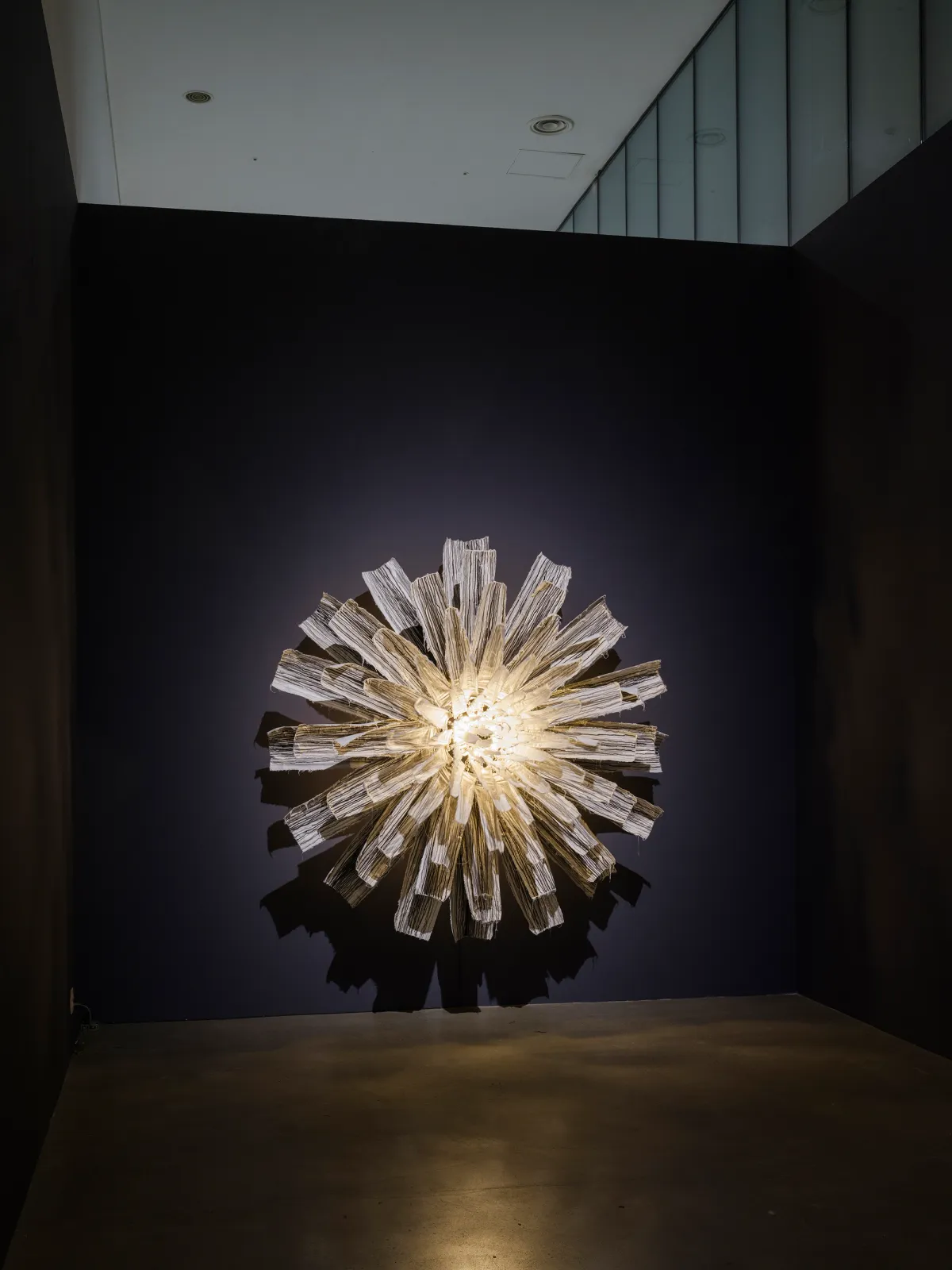 A wall-mounted art installation, reminiscent of a blooming flower, with a bright light at the center against a black wall.