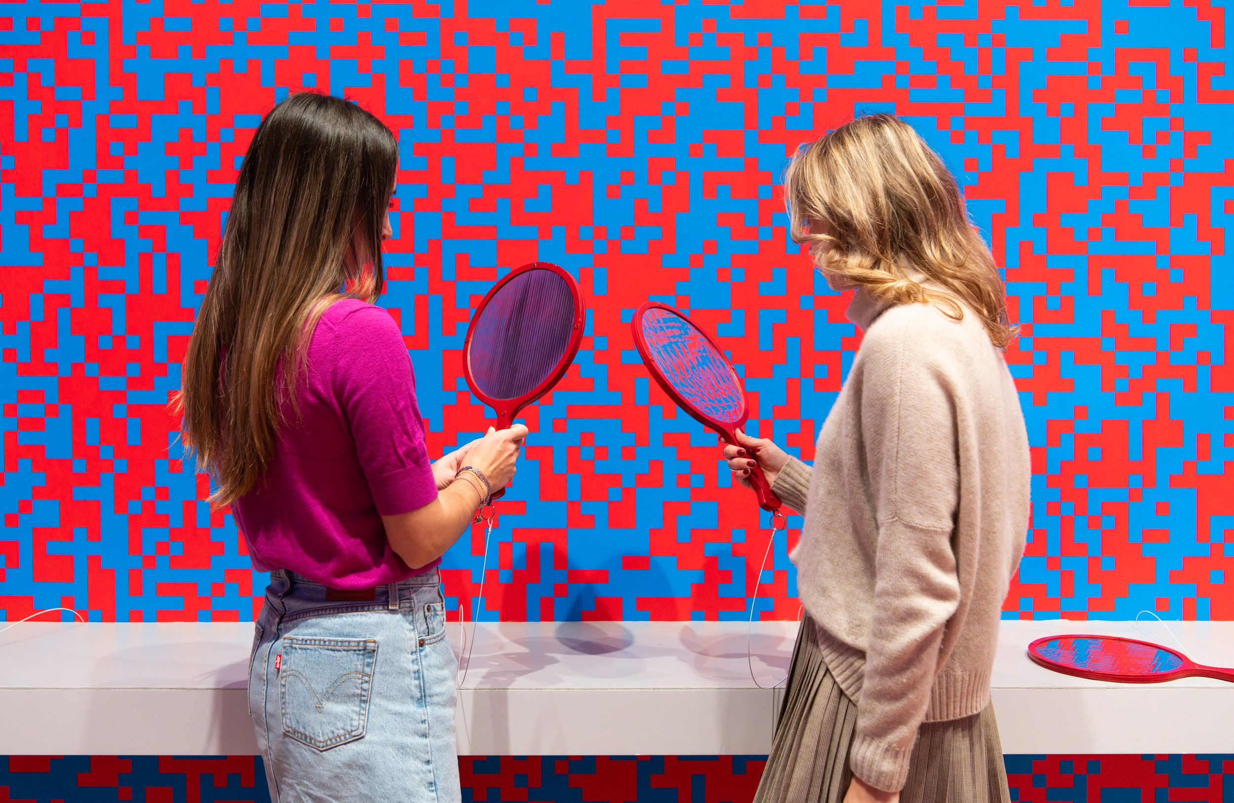 Two people stand in front of a vivid wall covered in a red and blue pixelated pattern, holding red electric fly swatters t...