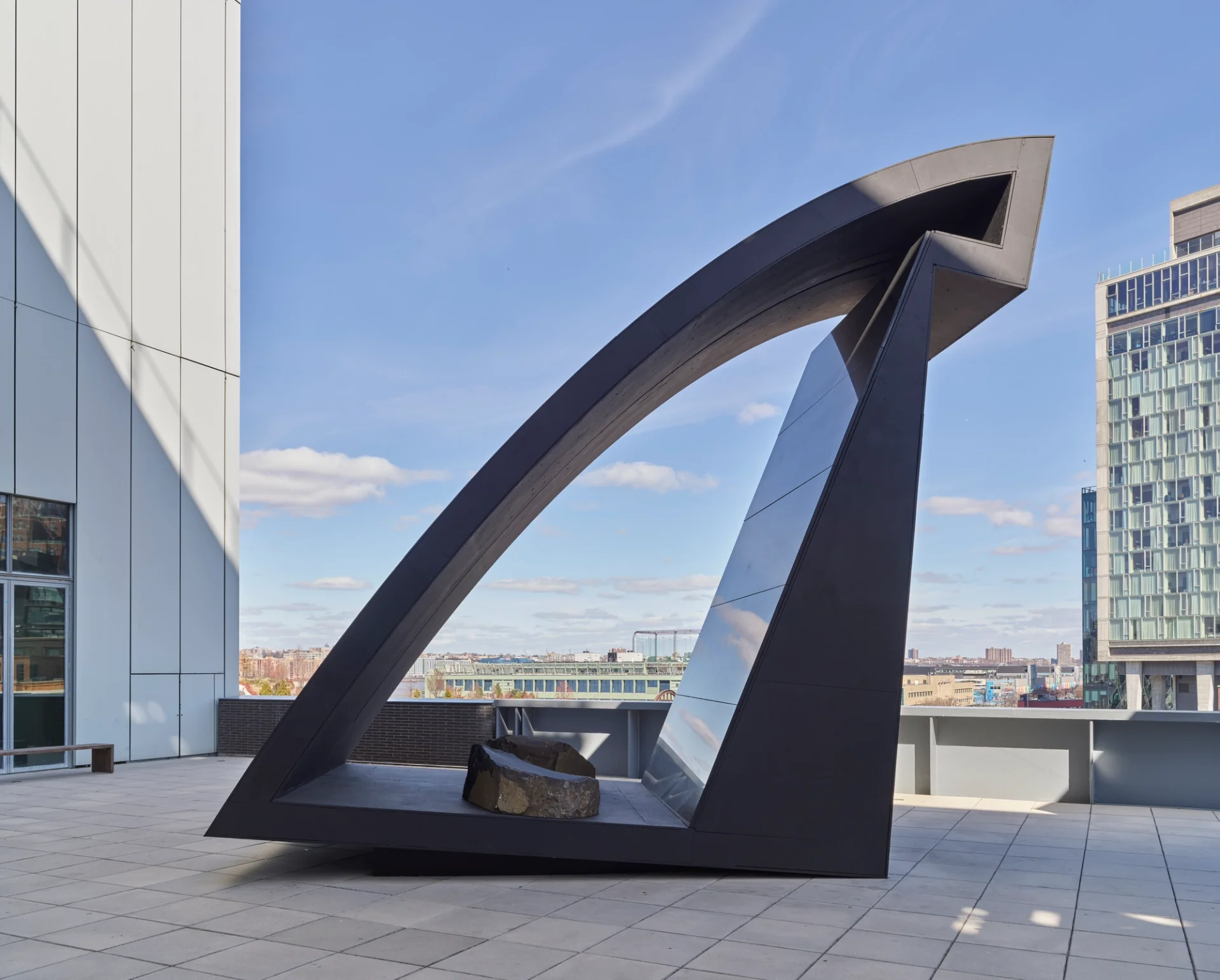 A sleek, black abstract outdoor sculpture featuring a curved arch and a reflective surface, positioned on a rooftop terrace with a cityscape in the background.