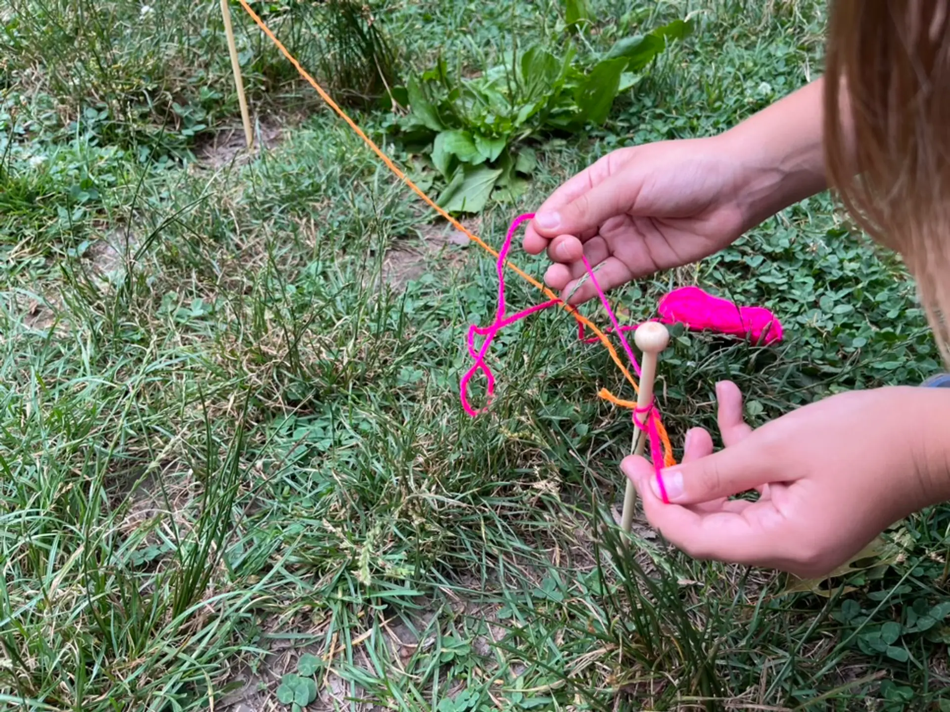 Close-up of hands tying pink yarn around wooden pegs on a grassy area.