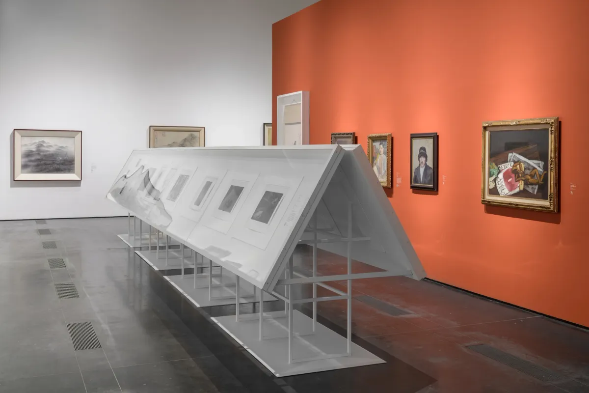 An art gallery room featuring various pieces displayed on metallic stands, with white frames, against an orange exhibition wall.
