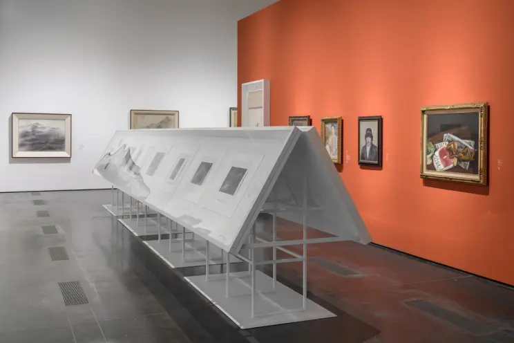 An art gallery room featuring various pieces displayed on metallic stands, with white frames, against an orange exhibition...