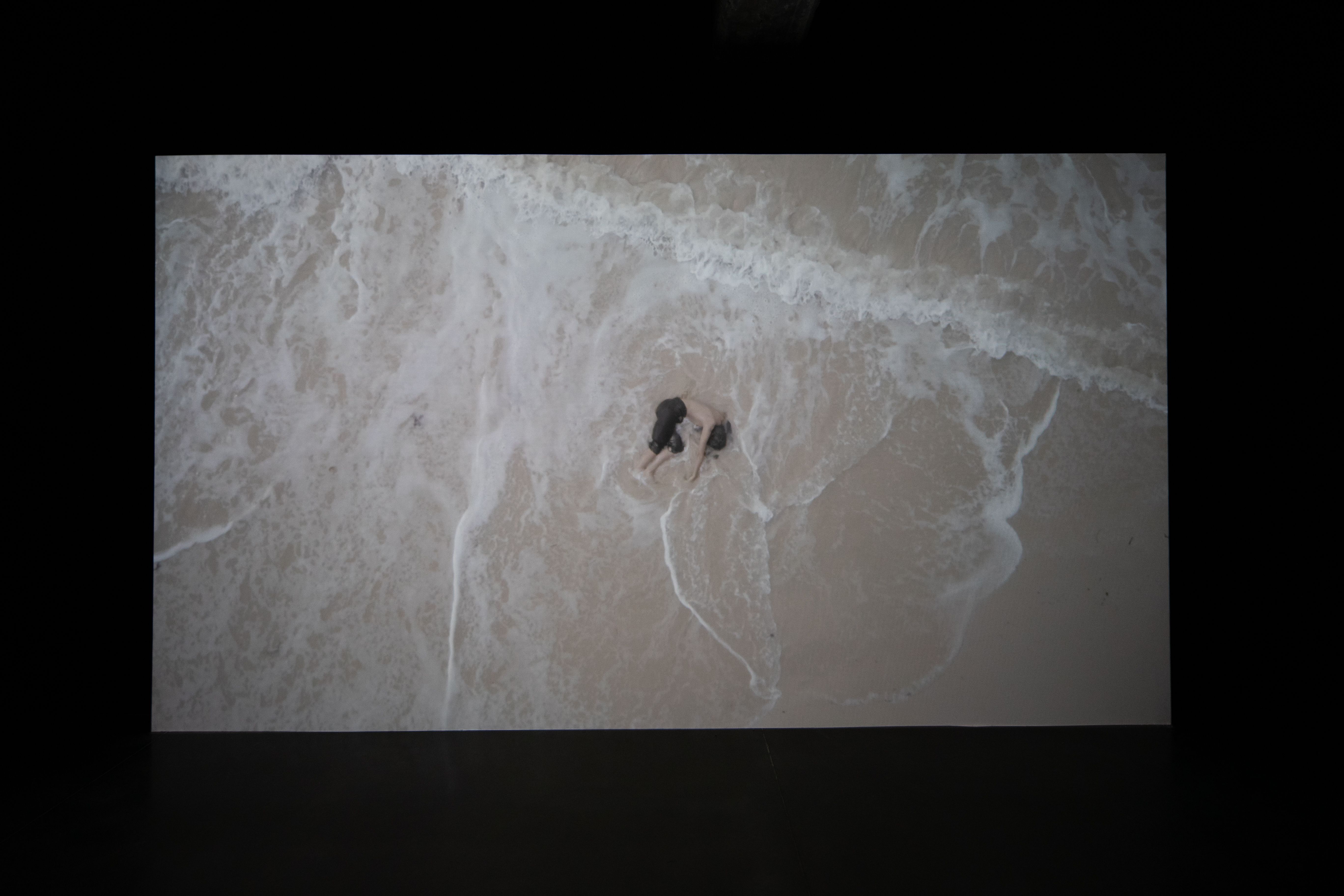 An aerial shot captures an individual curled up in the fetal position on a sandy beach, partially submerged in the shallow...
