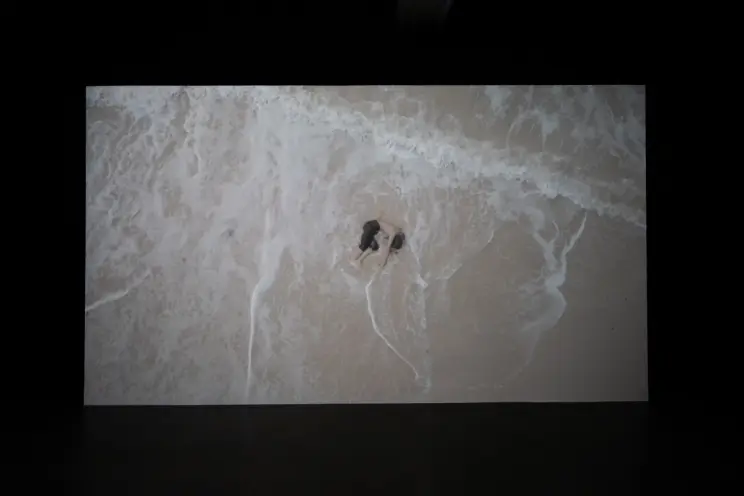 An aerial shot captures an individual curled up in the fetal position on a sandy beach, partially submerged in the shallow...