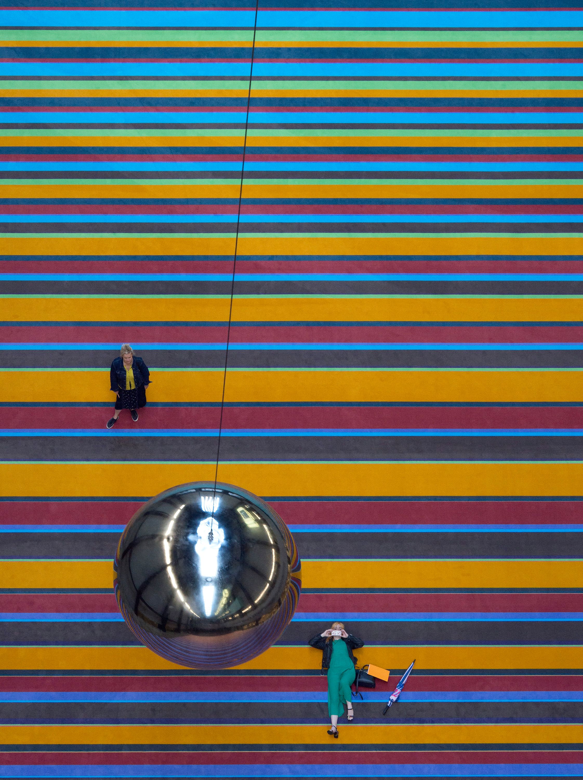 An overhead view of a person standing and looking up at a shiny sphere attached to a multicolored line on the floor, while...