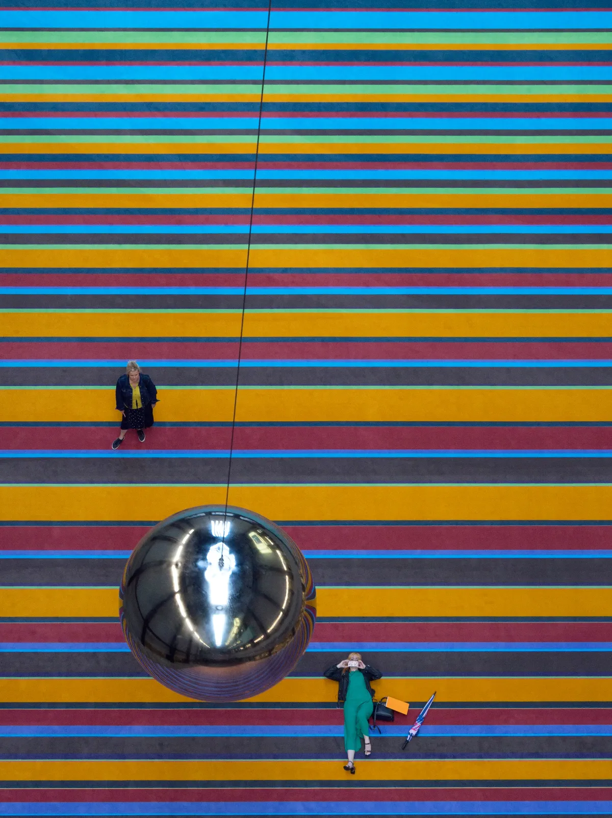 An overhead view of a person standing and looking up at a shiny sphere attached to a multicolored line on the floor, while another person lays nearby holding cake.