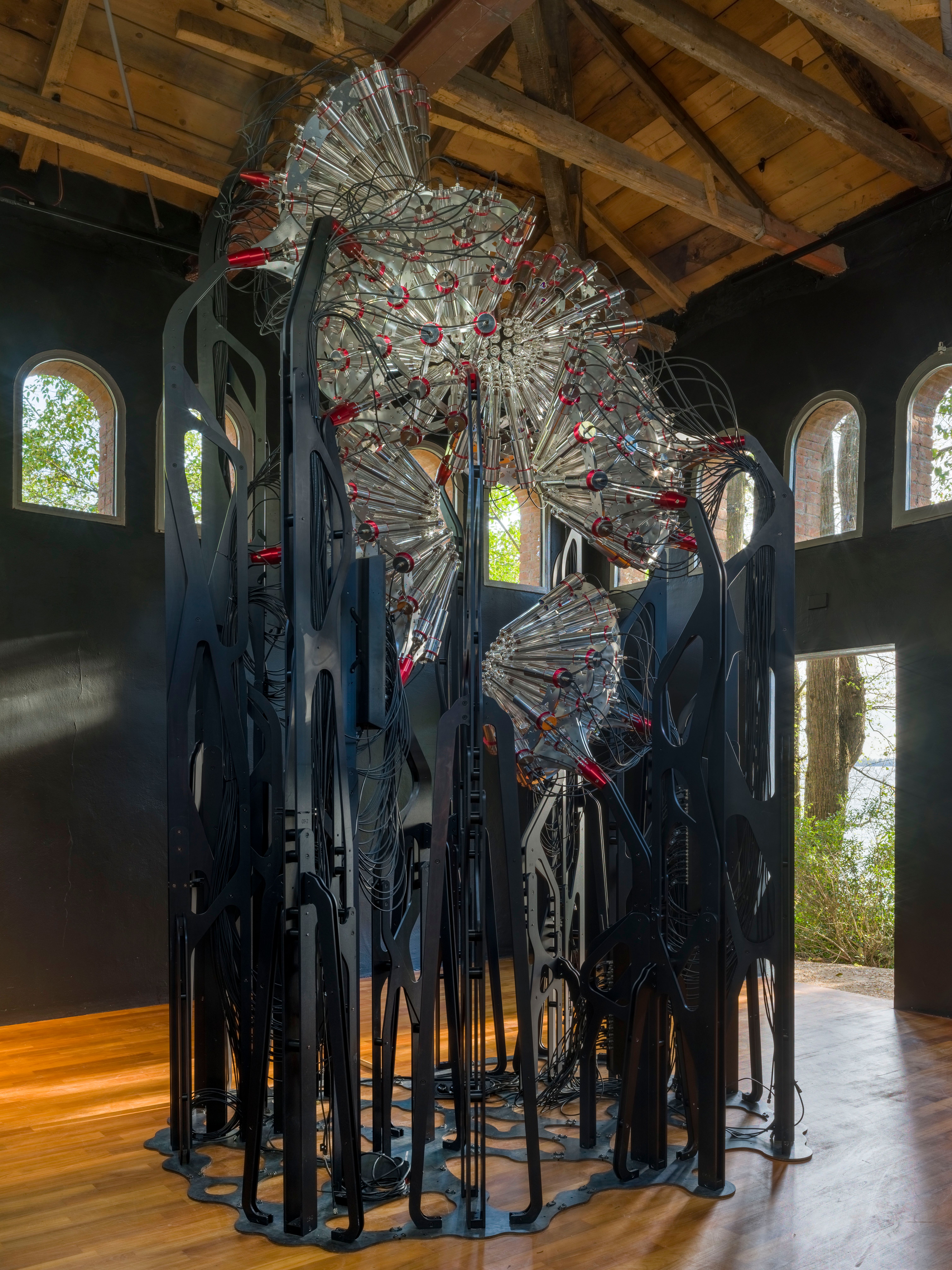 A metal and glass installation showcased in a room with dark walls, windows revealing surrounding nature.