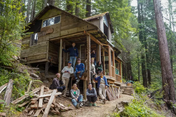 Group of people posing on the porch of a rustic cabin in the woods.