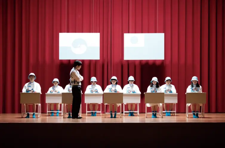 A theater curtain provides a backdrop for an adult, presumably a teacher, instructing students seated in a single row of d...