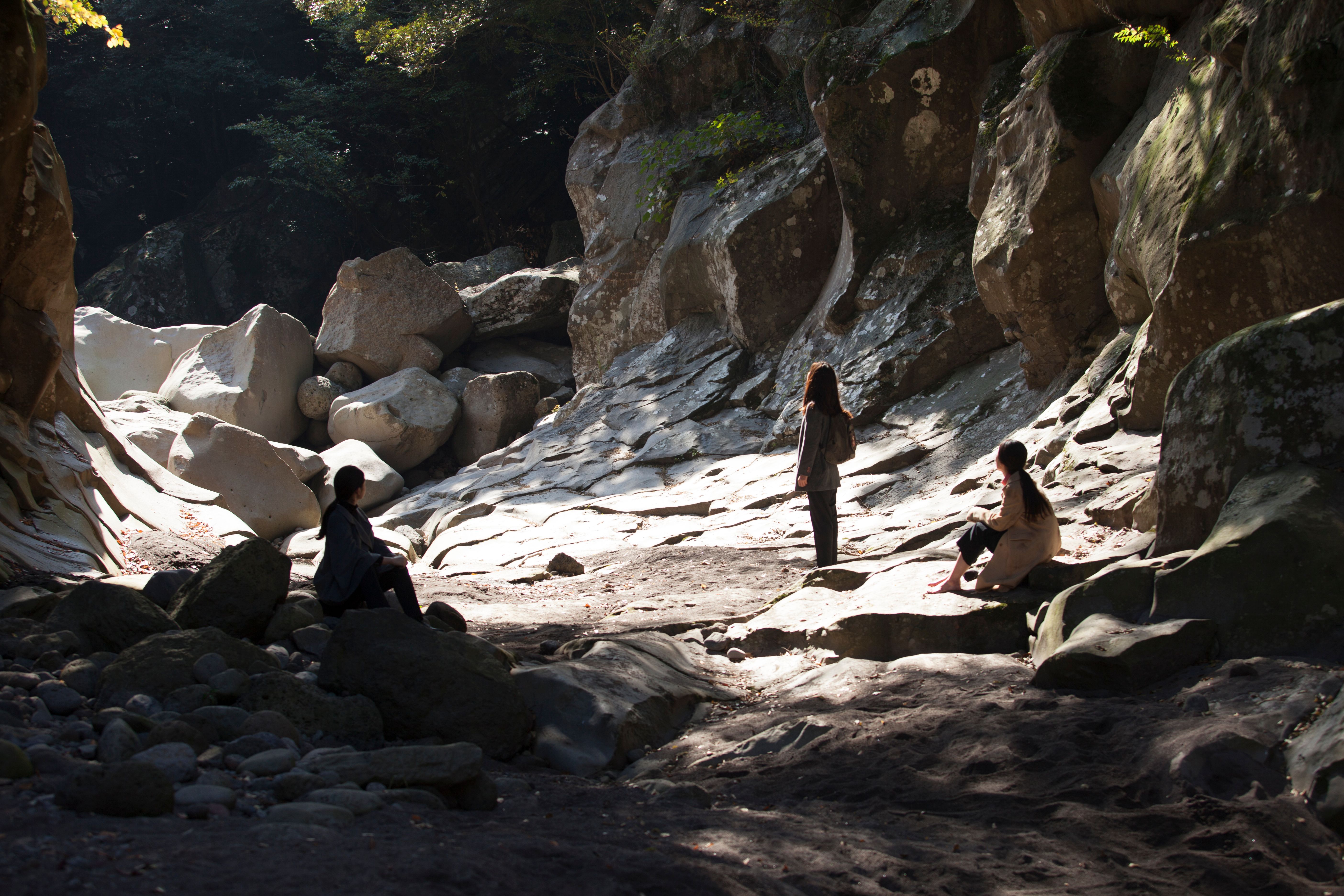 Three individuals stand with their backs to the camera, nestled between rugged rocks, as they gaze out at a vast, natural ...