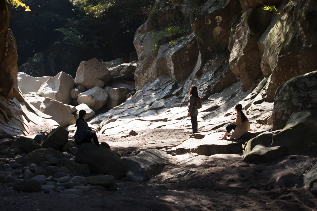 Three individuals stand with their backs to the camera, nestled between rugged rocks, as they gaze out at a vast, natural landscape in the distance.