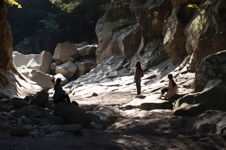 Three individuals stand with their backs to the camera, nestled between rugged rocks, as they gaze out at a vast, natural ...