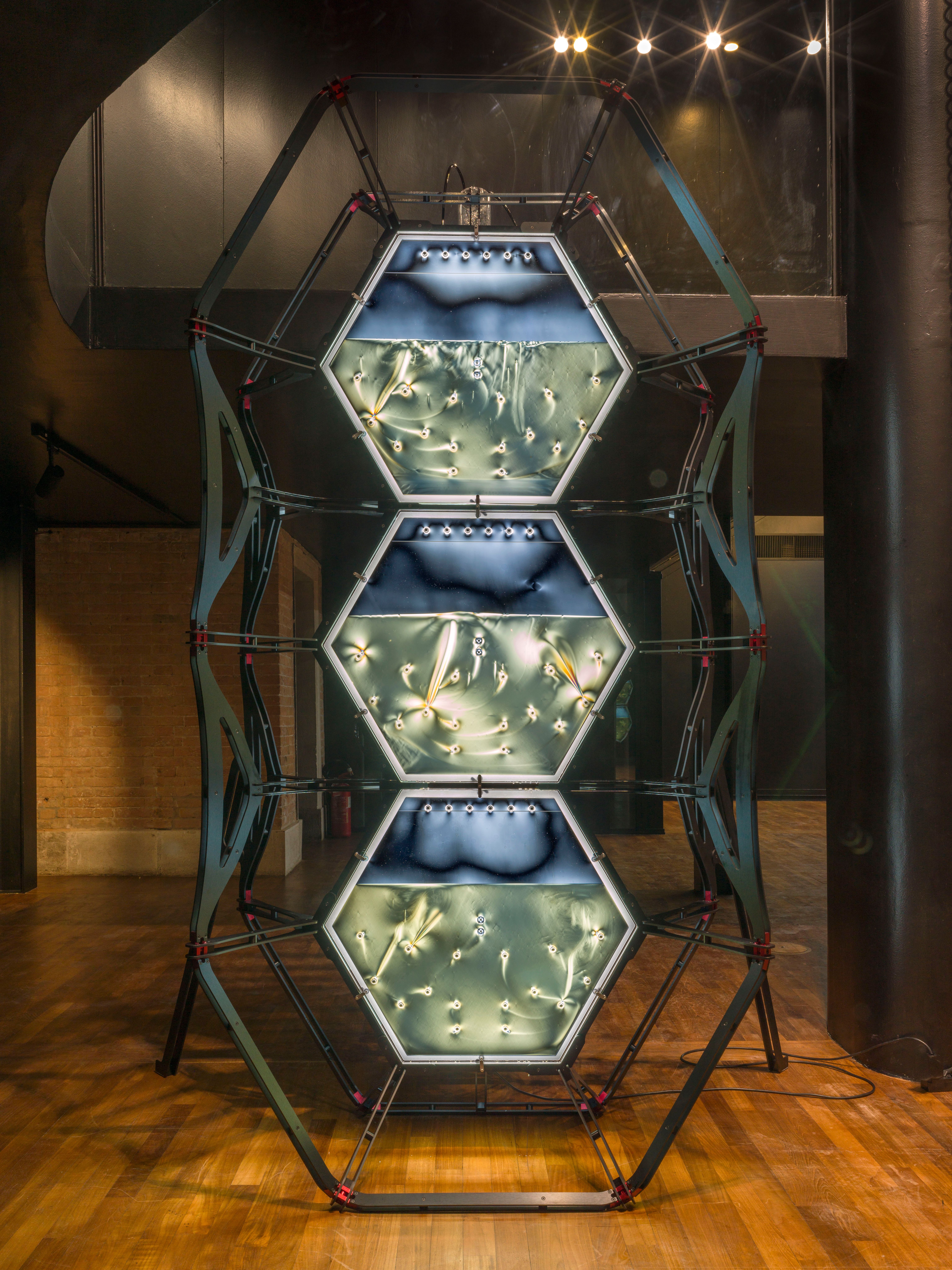 An industrial metallic installation consisting of three vertically stacked blue-lit hexagon shapes, resembling semi-filled...