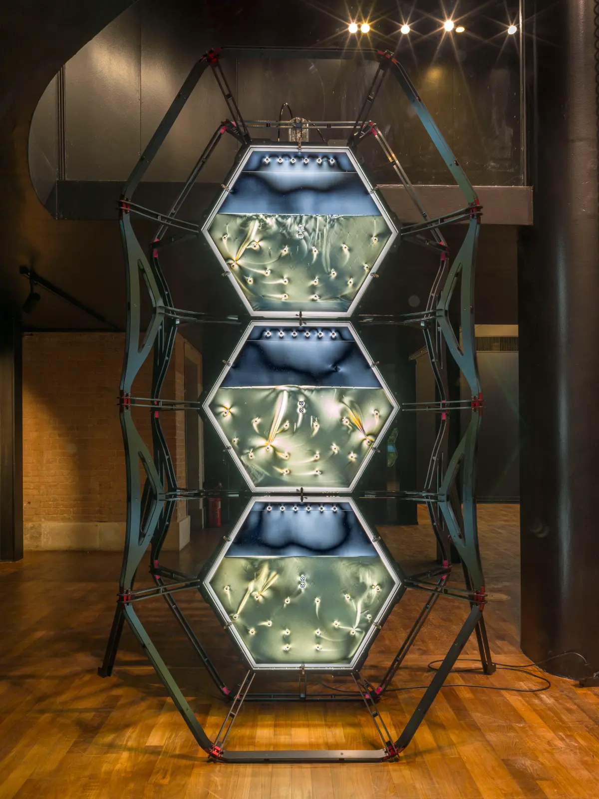 An industrial metallic installation consisting of three vertically stacked blue-lit hexagon shapes, resembling semi-filled aquariums, displayed under visual effect lighting.