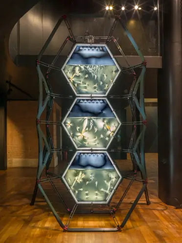 An industrial metallic installation consisting of three vertically stacked blue-lit hexagon shapes, resembling semi-filled...