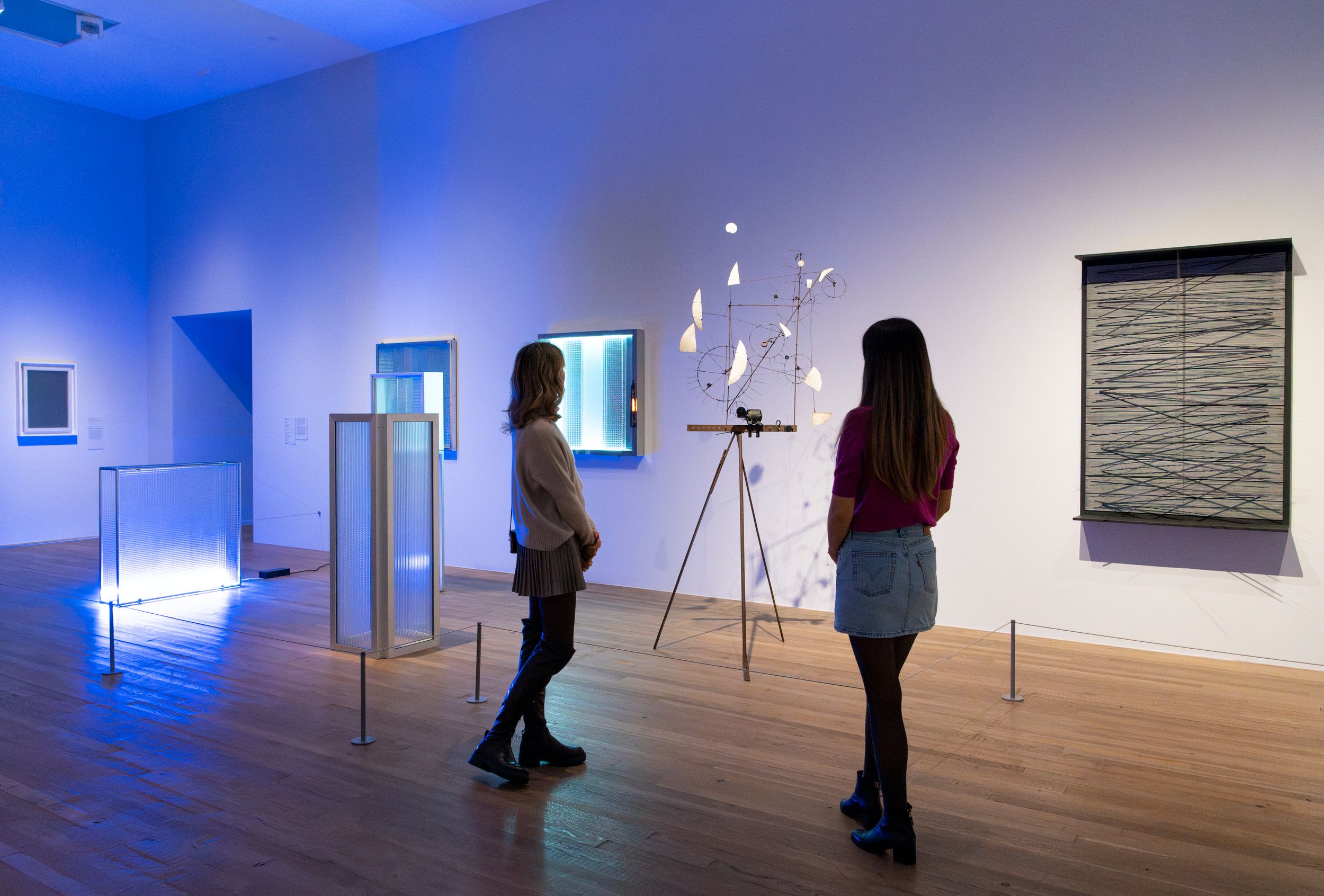 Two women stand in a brightly lit contemporary art gallery with wooden floors and blue ambient lighting. They observe a va...