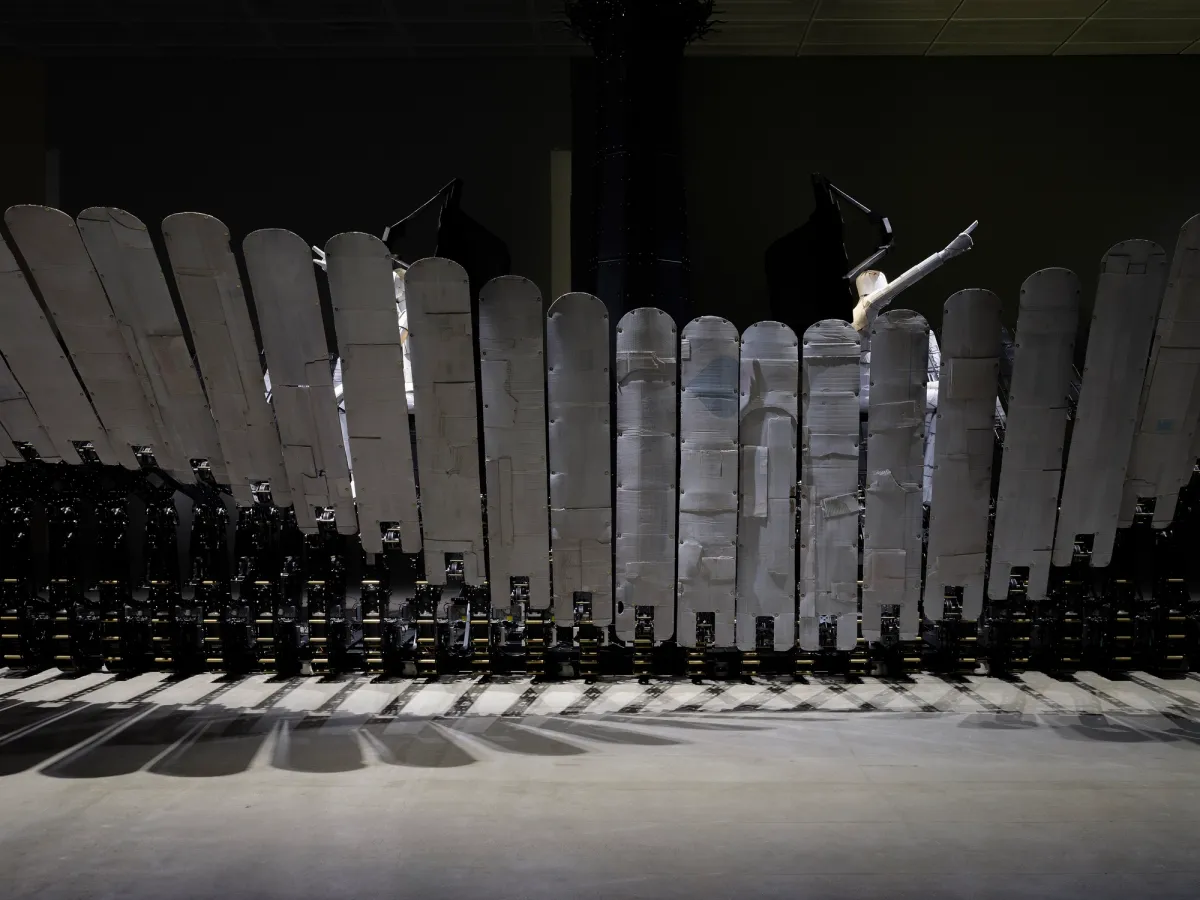 A large, wave-shaped fence installation made from recycled cardboard boxes, metallic materials, and electronic devices, displayed in a monochrome setting.