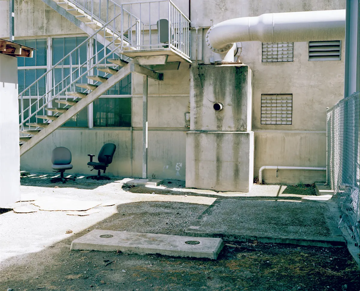 An old, unadorned concrete building with visible pipes on its exterior under an open sky. An outdoor stairwell and discarded office chairs are present.