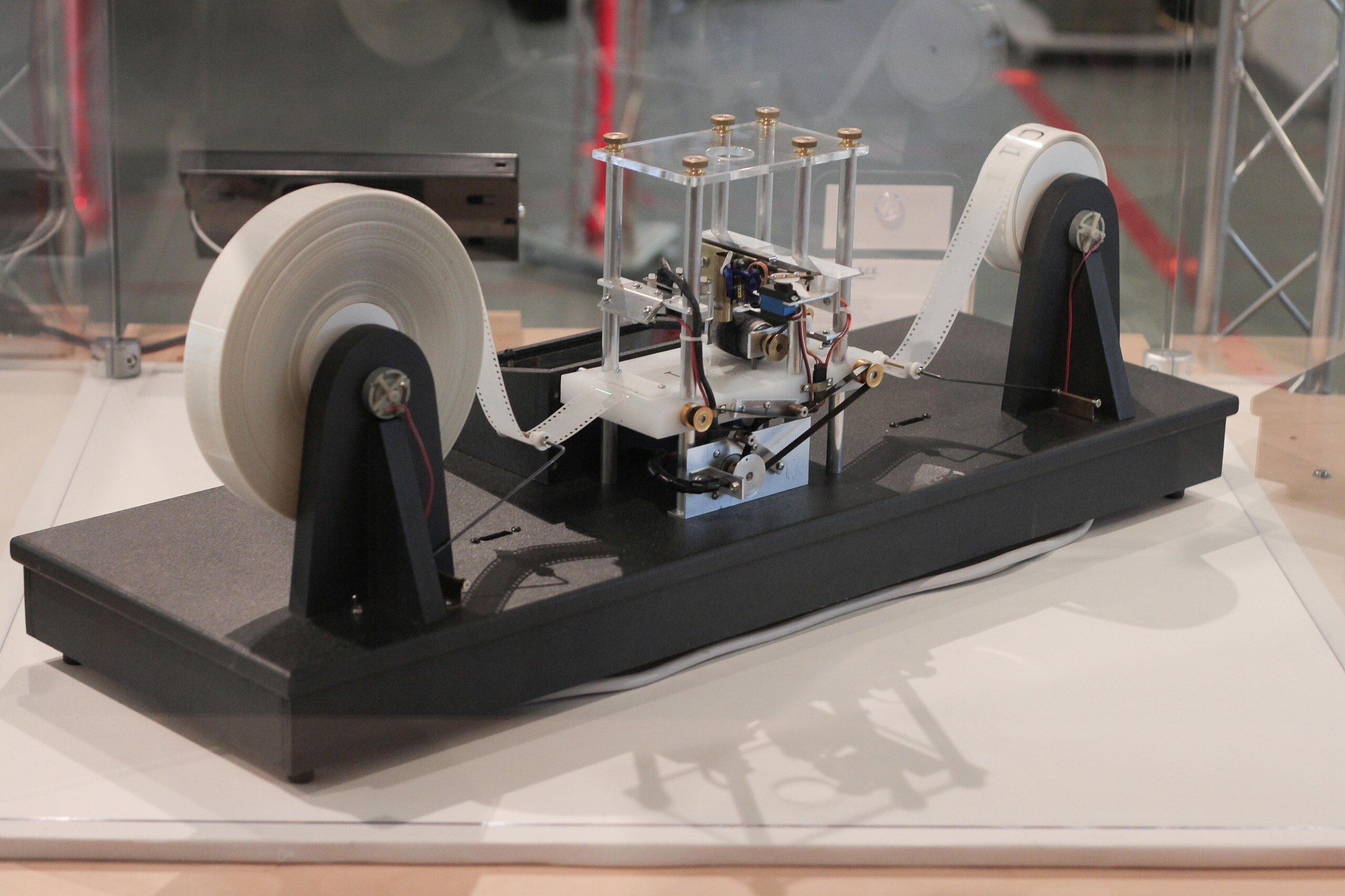 A physical model of a Turing Machine built with visible mechanical components: white tape reels on both ends, wiring, rollers, and a central mechanism housed in a transparent case. Displayed on a black base inside a museum or exhibit space.