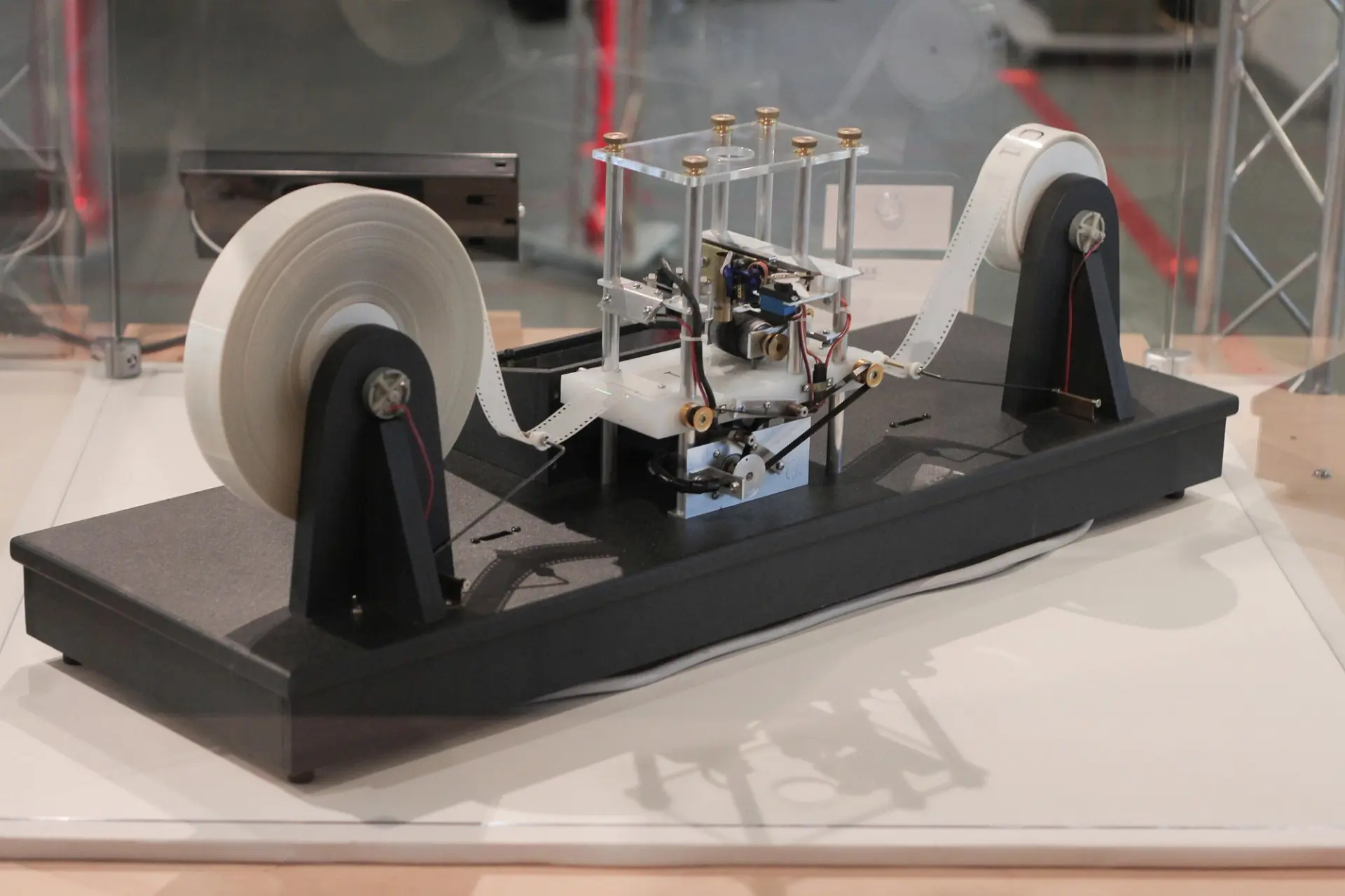 A physical model of a Turing Machine built with visible mechanical components: white tape reels on both ends, wiring, rollers, and a central mechanism housed in a transparent case. Displayed on a black base inside a museum or exhibit space.