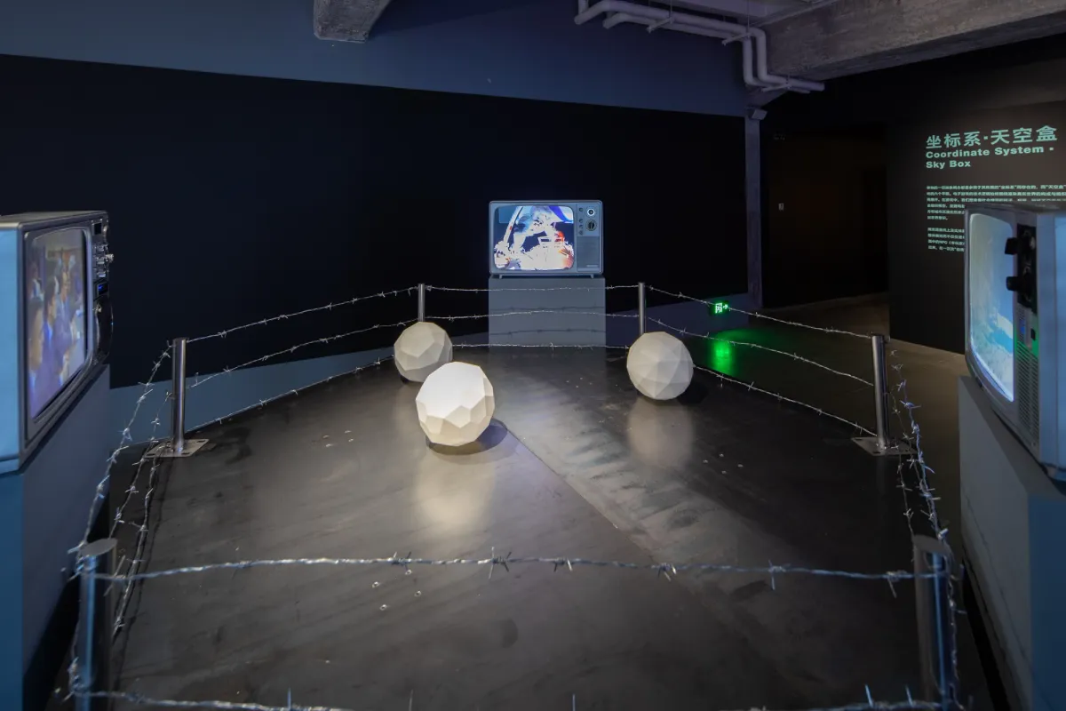 An art installation featuring three round white shapes situated within a battle ring made from barbed wire. On the periphery, three monitors provide additional context or details about this interactive exhibit.