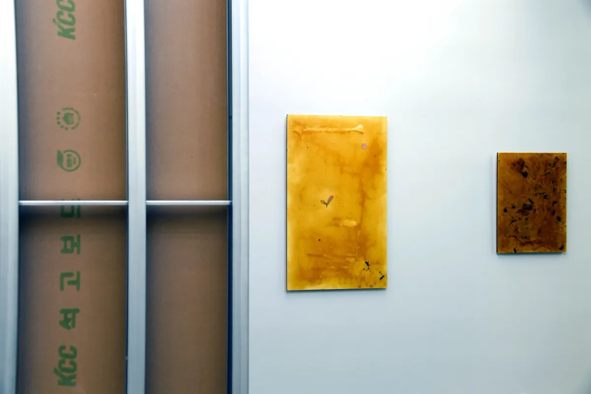 Three art pieces displayed on a wall: a large window frame installation with cardboard behind the glass, a medium-sized abstract canvas in yellow, and a smaller brown abstract canvas.