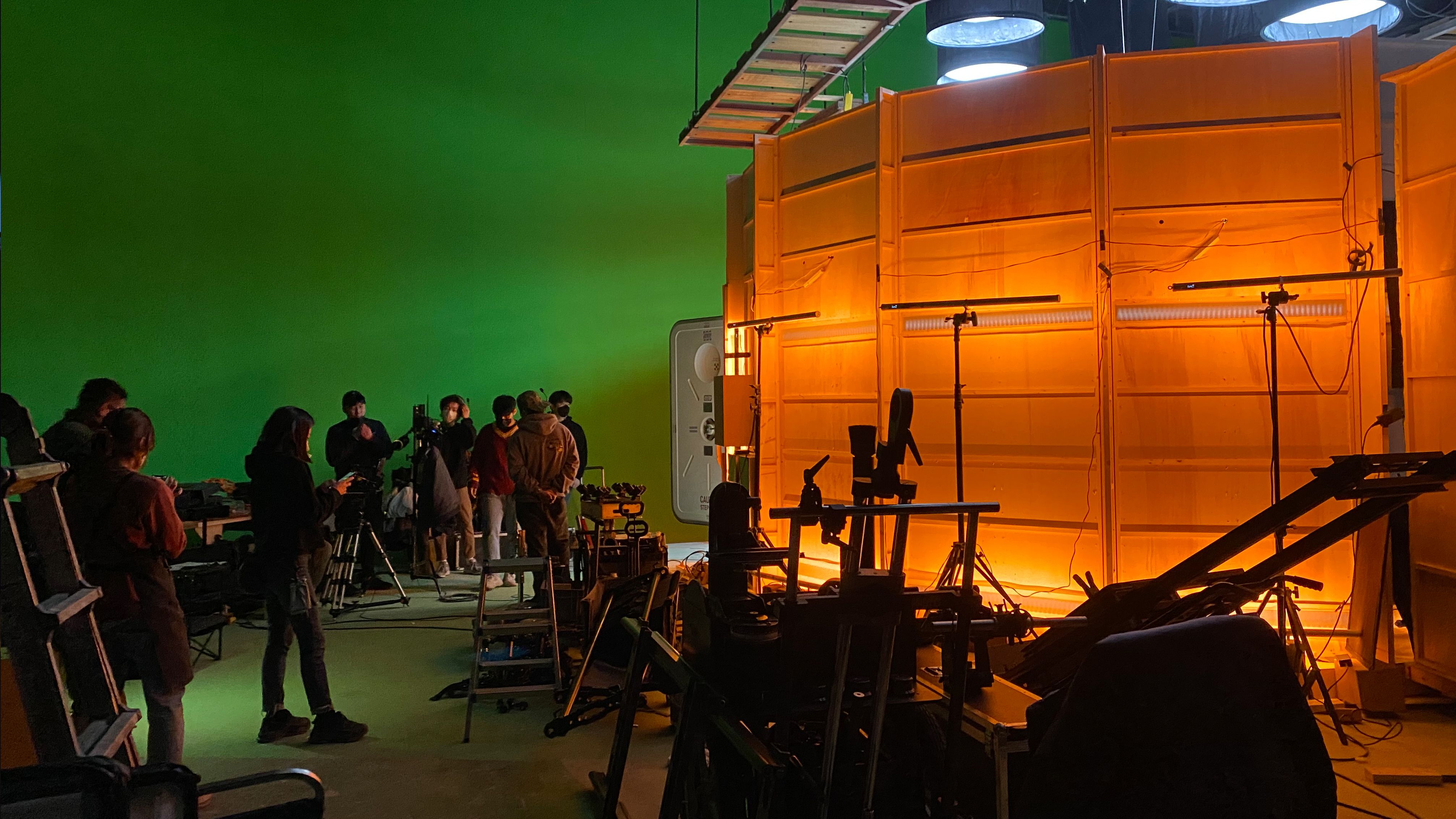 A dimly lit studio stage where professionals are working amongst a variety of equipment. An ambient, soft orange light ill...